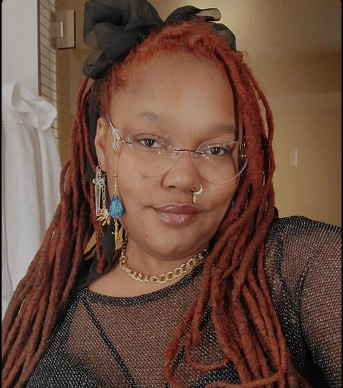 Bluu, a Black nonbinary baddie with long, red locs is smiling slightly at the camera. Fae is wearing heart shaped glasses, gold jewelry, and a net top.